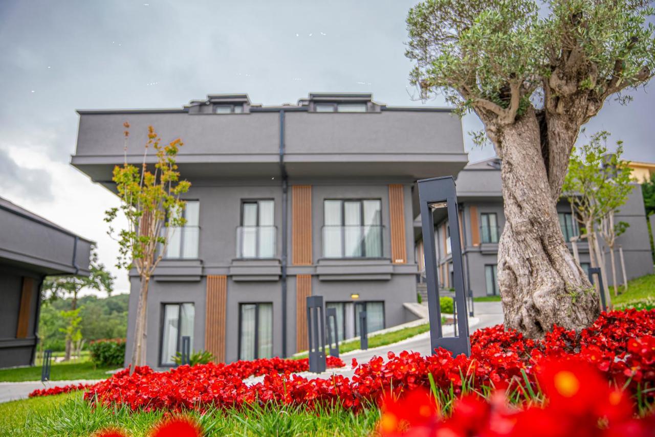 Menalo Hotel Premium Istanbul Airport Exterior foto The photo features a modern, two-story building set within a landscaped area. The building has a sleek design with large windows and a neutral color palette, primarily in shades of gray and brown. In the foreground, there's a vibrant display of red f