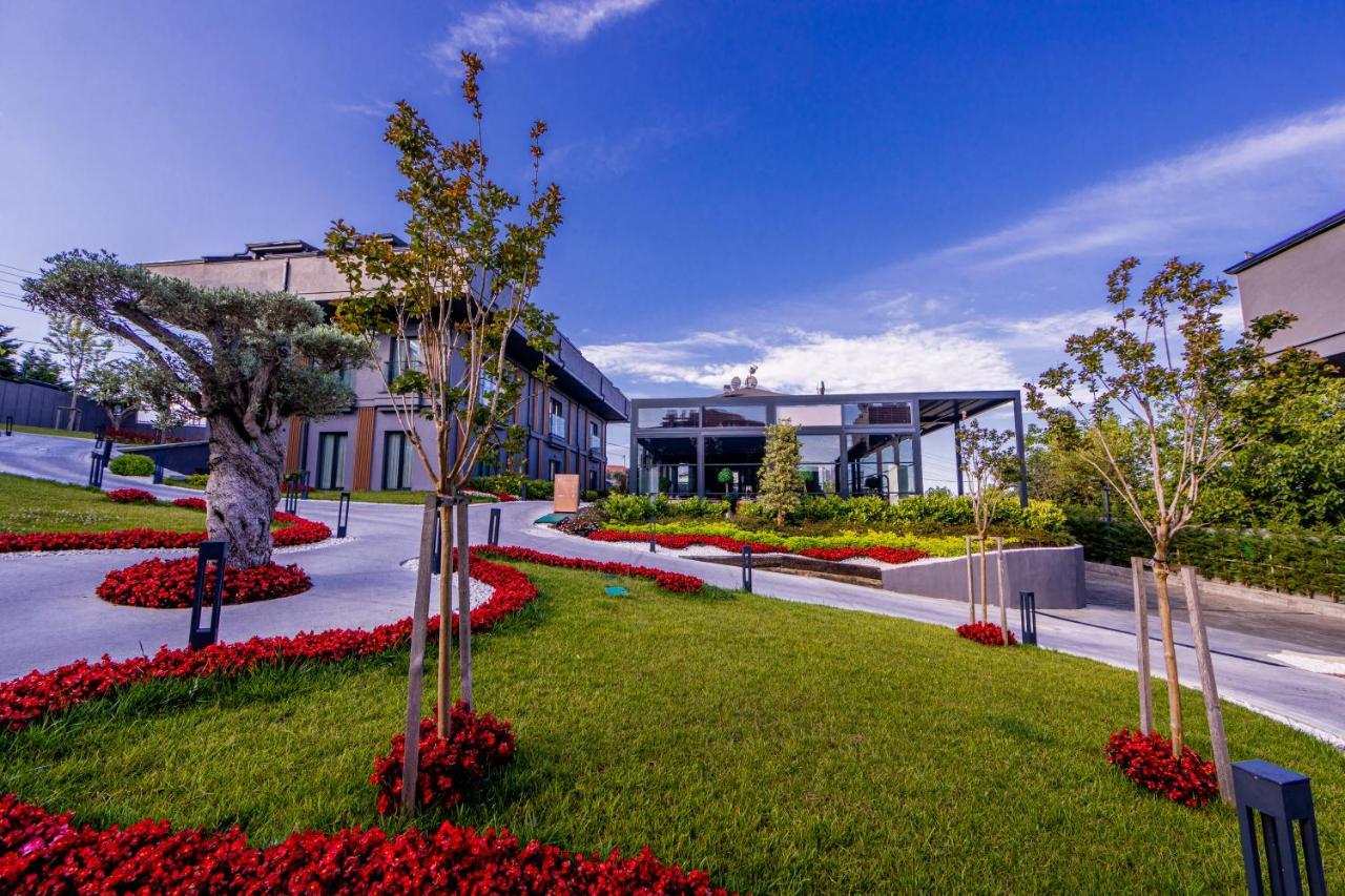 Menalo Hotel Premium Istanbul Airport Exterior foto The photo shows a landscaped outdoor area featuring a modern building surrounded by lush greenery. The foreground has a well-maintained lawn with vibrant red flower beds, and some small trees are planted along the pathway. The architecture appears co