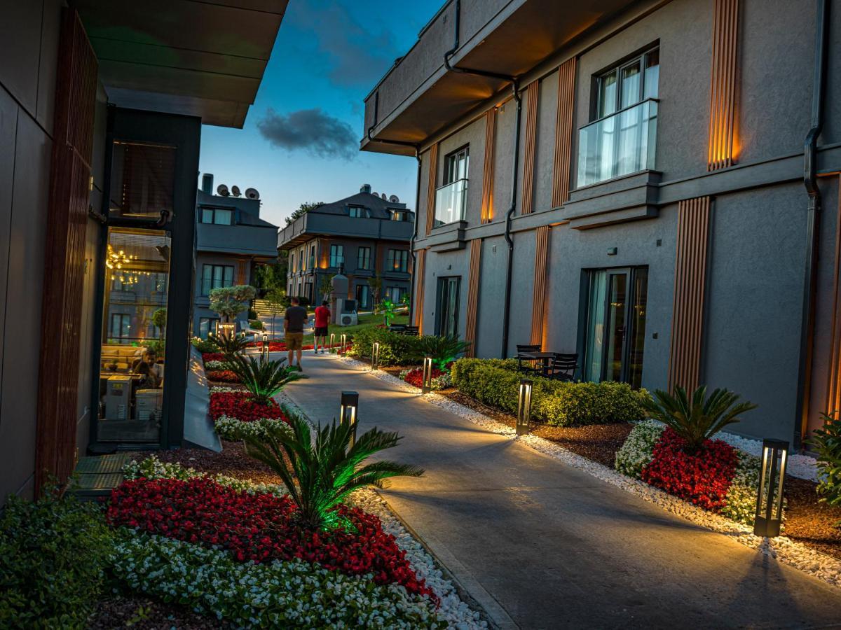 Menalo Hotel Premium Istanbul Airport Exterior foto The photo shows a pathway between modern buildings, illuminated by soft lighting. The path is lined with colorful flower beds featuring red and white blooms and small palm plants, adding to the aesthetic appeal. The buildings are designed with a mix 