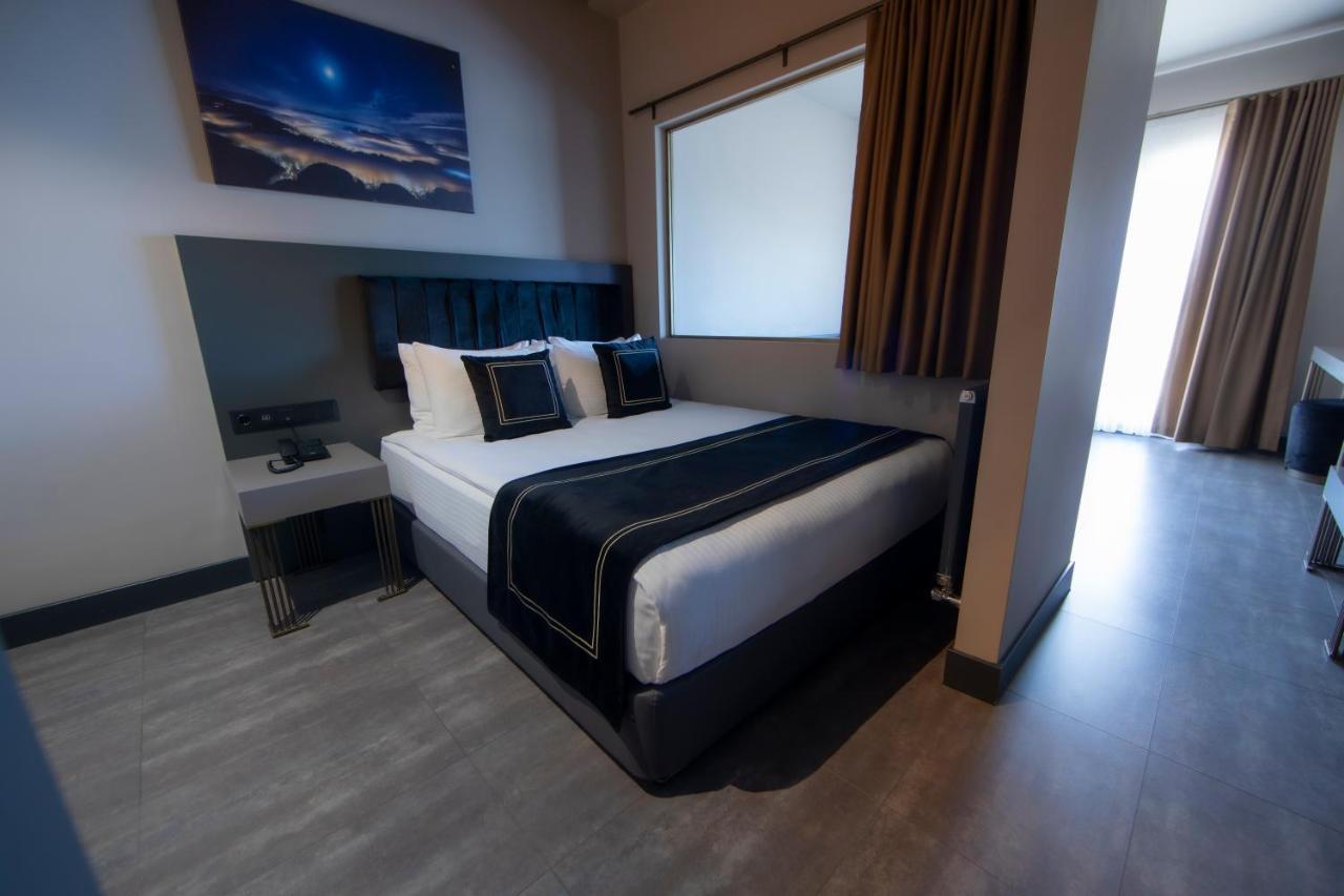 Menalo Hotel Premium Istanbul Airport Exterior foto The photo shows a modern hotel room featuring a double bed with black and white bedding. There are two decorative pillows on the bed, and behind it is a wall-mounted headboard. To the left of the bed, there's a small bedside table with a lamp. A larg
